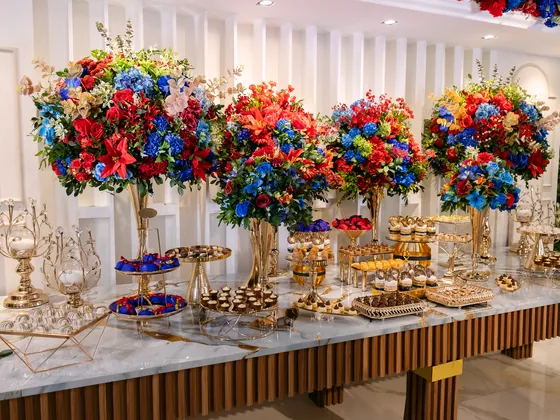 Mesa de doces com quatro arranjos altos de flores vermelhas e azuis